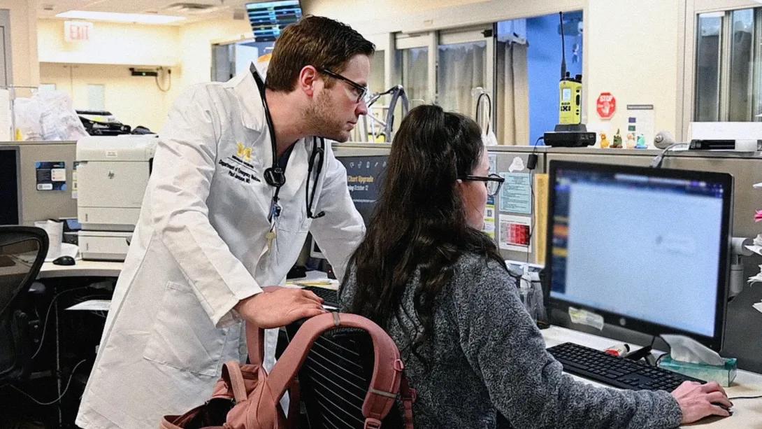 clinicains at work station looking at the same computer one in a white coat one in grey sleeves facing computer