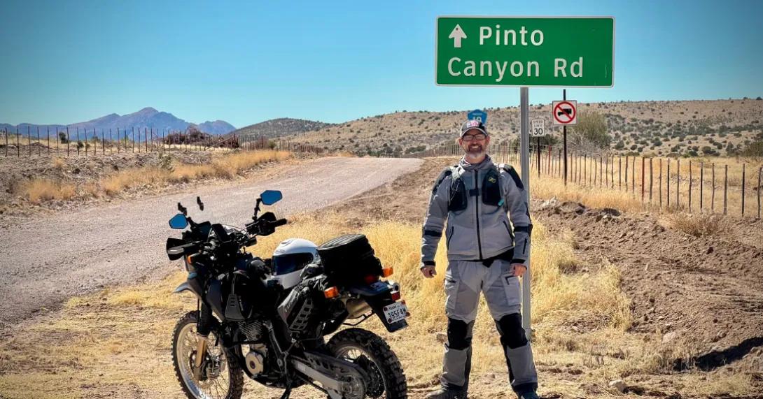 A man stands next to a motorcycle in front of a sign that says "pinto canyon"