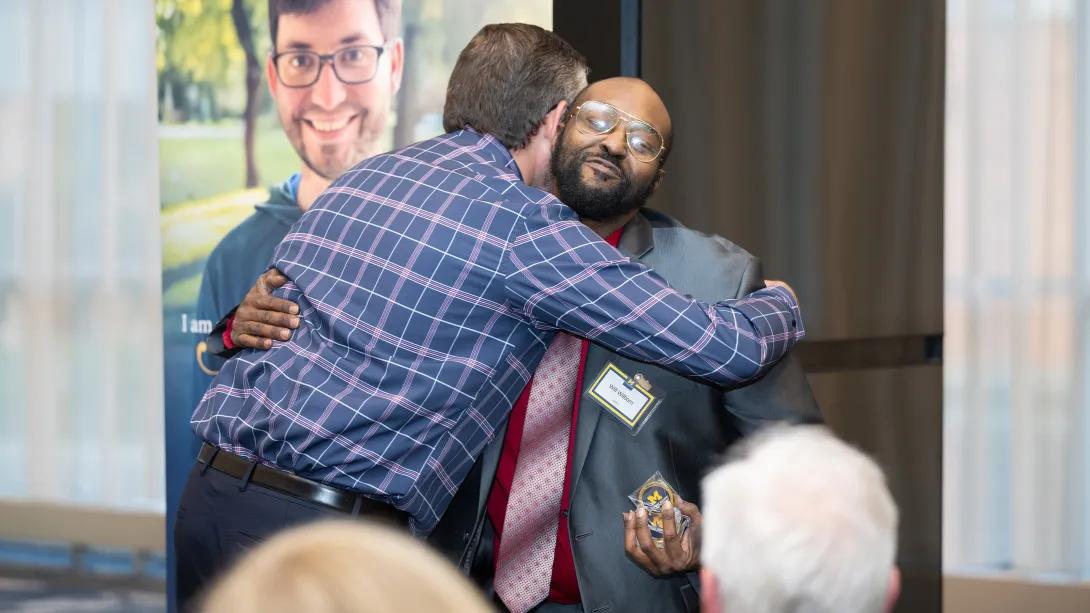A man in a suit with a tie hugs another man in a plaid shirt