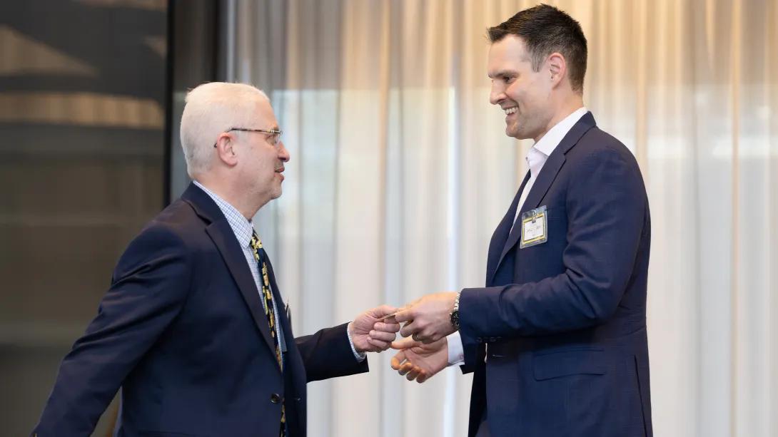 A man in a suit hands another man in a suit a token with the University of Michigan logo on it