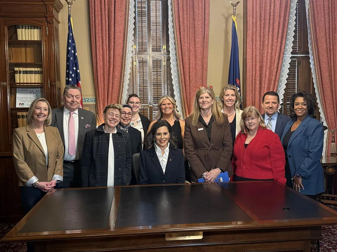 Laura Bonnell, Samya Nasr, and other advocates stand around Governor Whitmer, who is seated at her desk.