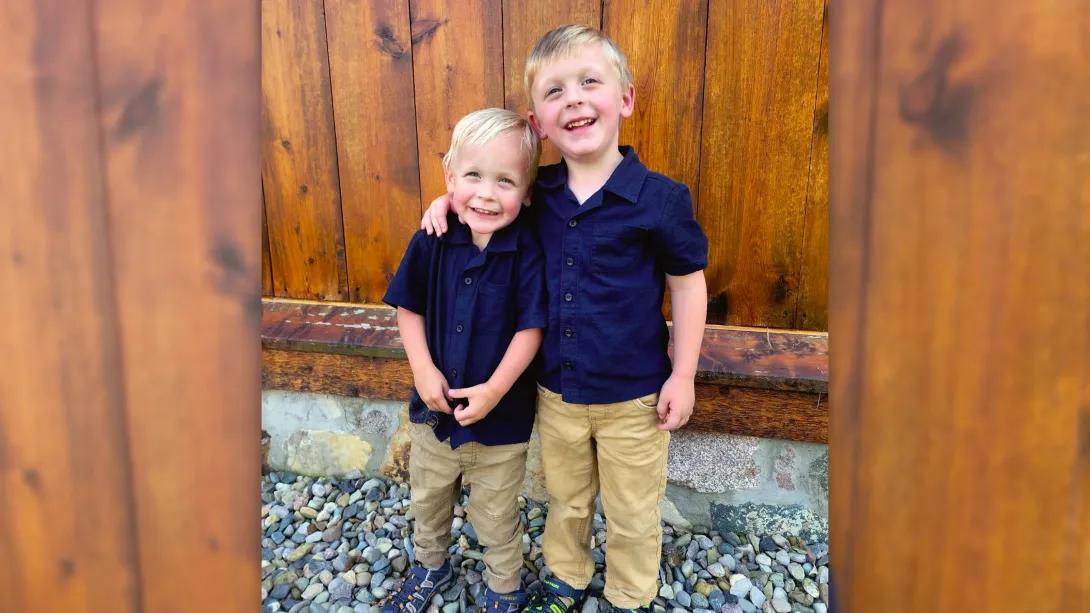 Two young boys stand in front of wood wall, with bigger one's arm around smaller one. Wearing blue shirts and khaki pants