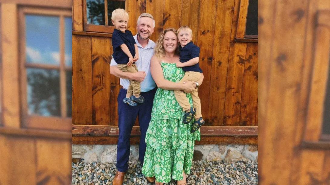 Family smiling in front of wood wall, mom, dad each hold a young blond boy. Mom wears green dress, dad blue shirt and pants and boys in navy blue shirts with khaki pants