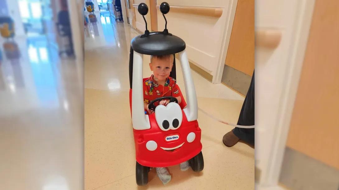little boy in red pajamas drives a toy car down a hospital hallway