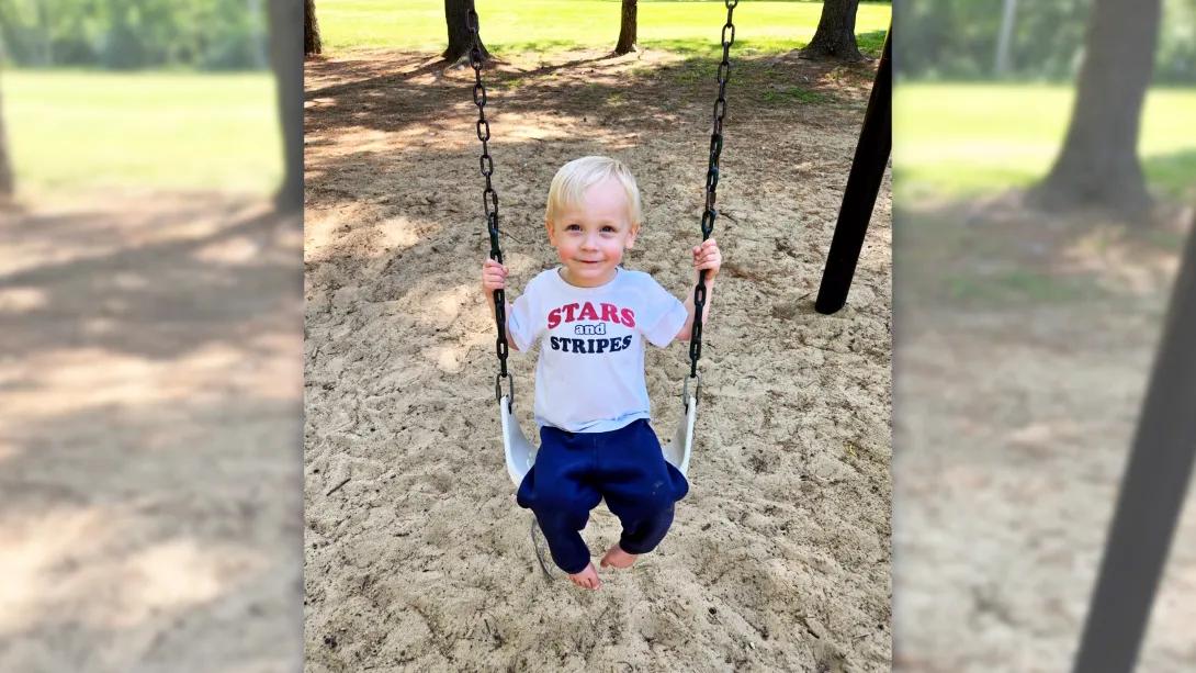 Blond young boy sits on a swing in a sandy playground, wearing shirt that says stars and stripes