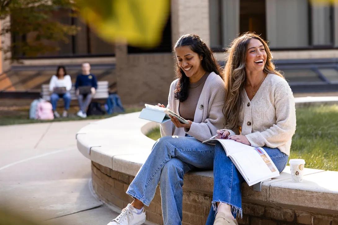 Two medical students talk and laugh as they take a break from studying. They are seated on a low wall along a sidewalk and grassy area.