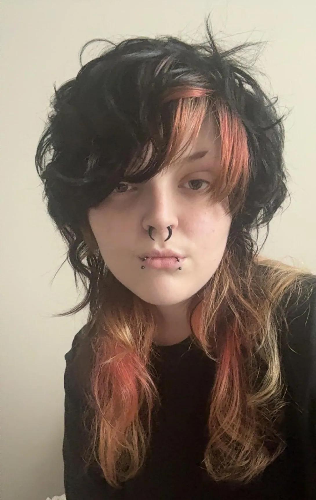 A photo of Leo Saunders, who describes themself as having vibrant red and black hair with hints of purple, pale skin, green eyes, and facial piercings.