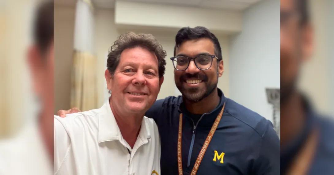 Two people are taking a selfie together indoors. They are both smiling. One person is wearing a white shirt, and the other is wearing a dark jacket with a yellow "M" logo on it.