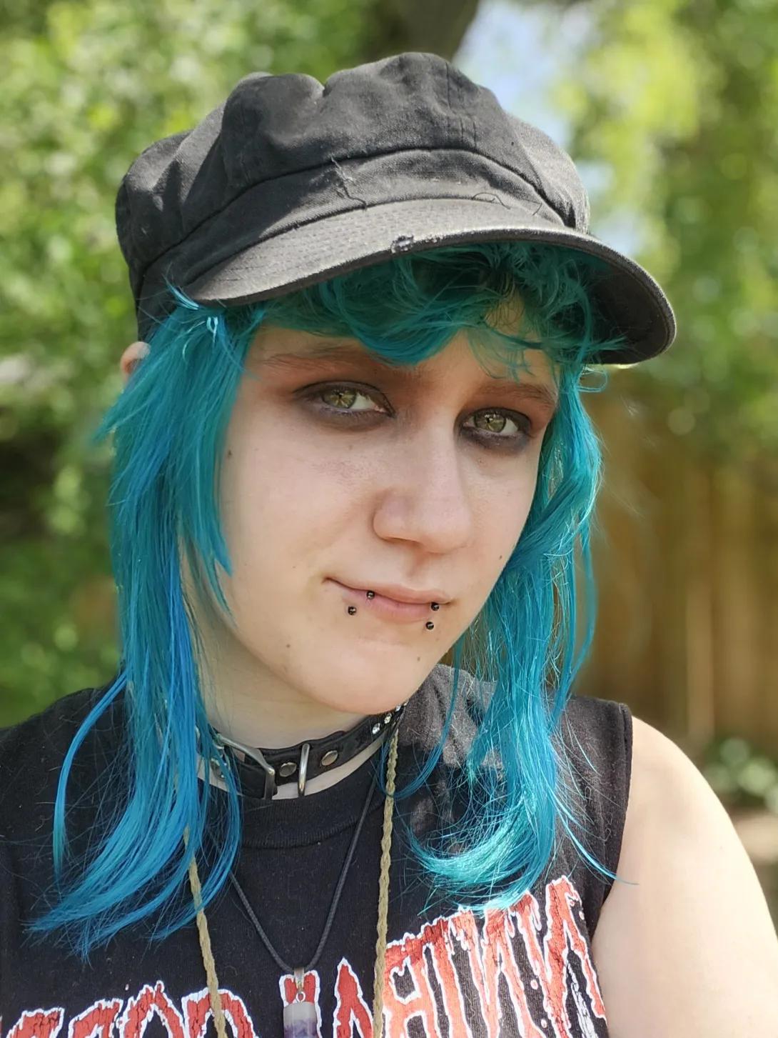 A photo of Nick Miltgen in front of a wooded background, wearing a black hat, choker, and sleeveless band shirt.