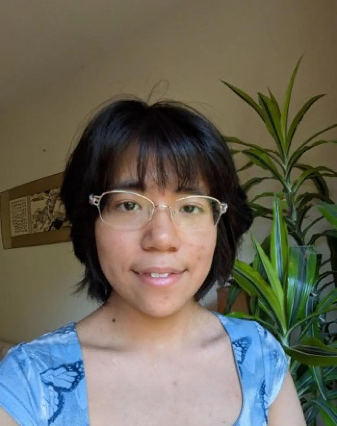 A photo of Suhan Zhu, who describes herself as having black hair, tan skin, and dark brown eyes. In the background is a large plant, and Suhan is wearing a blue butterfly top.